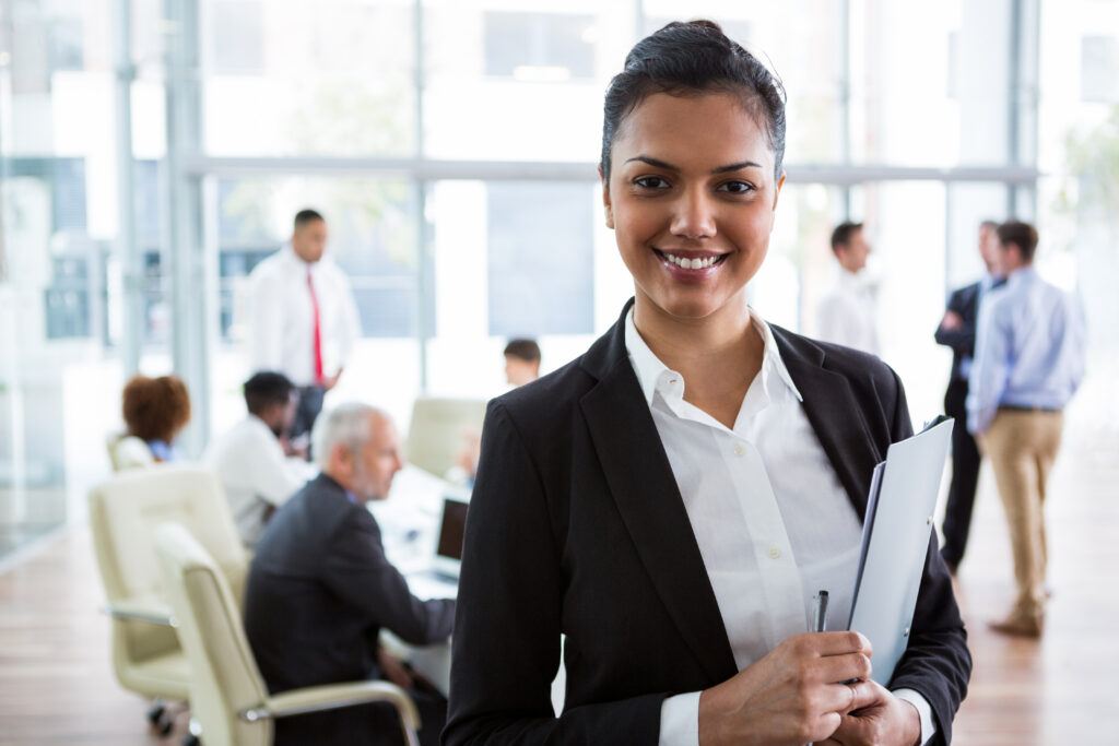 A young business professional standing confidently, smiling as her team discusses in the background.